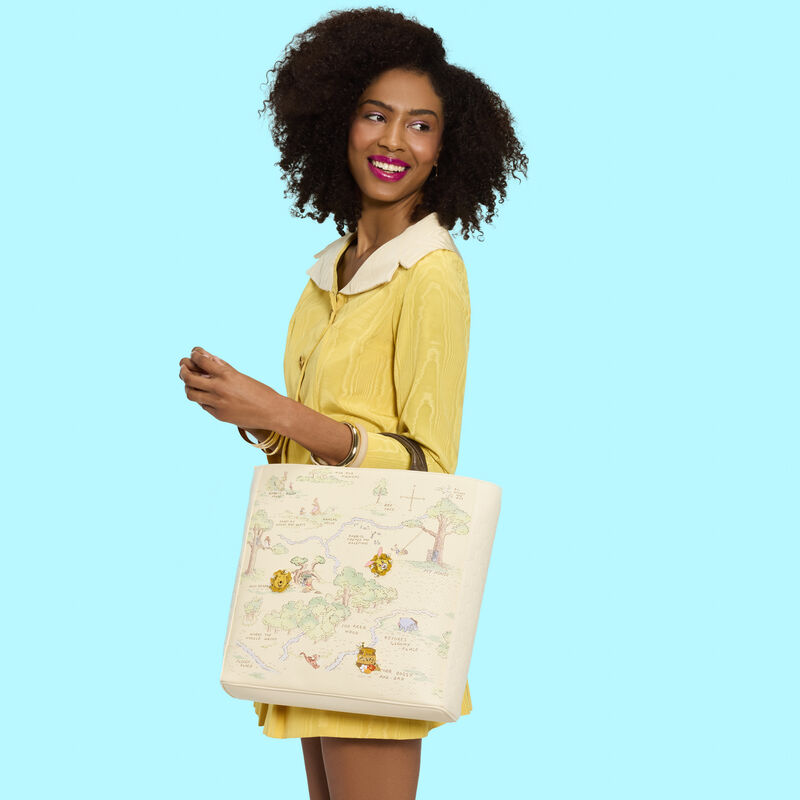 Woman wearing a yellow dress, facing sideways to camera, she smiles and looks over a shoulder, wearing the Loungefly Winnie the Pooh tote over one arm. 